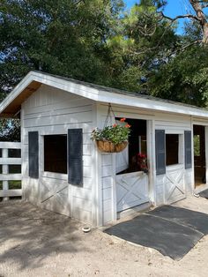 a small white shed with black shutters and a potted plant hanging from the door