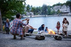 a group of people sitting around a campfire next to a body of water and trees