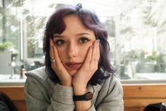 a woman sitting at a table with her hands on her face and looking into the camera