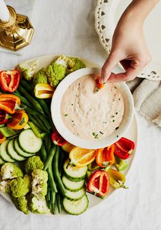 a person dipping sauce into a bowl of dip surrounded by veggies on a plate