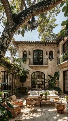 an outdoor living area with couches and tables under a tree in front of a house