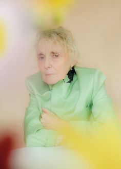 an older woman wearing a green jacket and black tie sitting in front of a yellow background