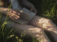 a woman laying in the grass reading a book with her legs spread out and holding an open book