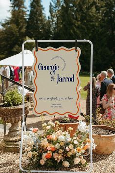 a sign that says george and jarrod is surrounded by potted plants in front of people