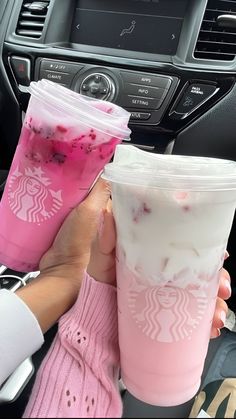 two people holding up cups with drinks in them on the dashboard of a car,