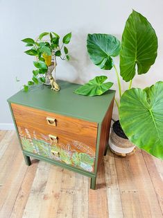 a green and wooden chest sitting on top of a hard wood floor next to a potted plant