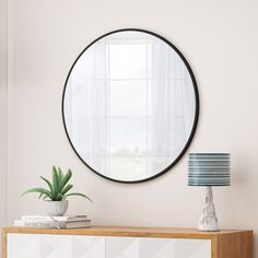 a round mirror hanging on the wall above a dresser with a plant and lamp next to it