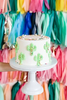 a white cake with green frosting and cactus decorations on it, sitting in front of colorful streamers