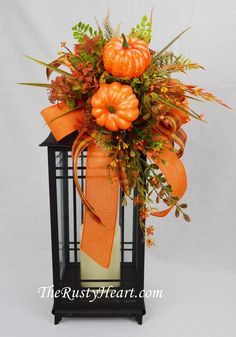 an arrangement of pumpkins and greenery in a black lantern with orange ribbon on it