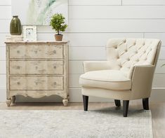 a white chair sitting in front of a dresser next to a vase on top of a table