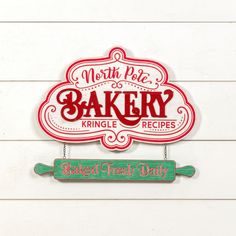 a red and green sign hanging from the side of a white wooden wall that reads, north pole bakery kind of knickle recipes