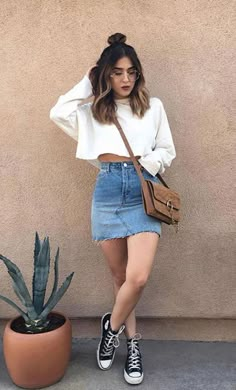 a woman standing in front of a wall wearing a white top and denim skirt with black converse