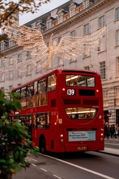 a red double decker bus driving down a street next to tall buildings with christmas lights on them