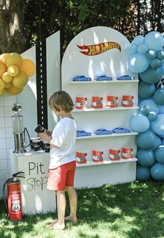 a little boy standing in front of a stand with hot wheels on it and lots of balloons