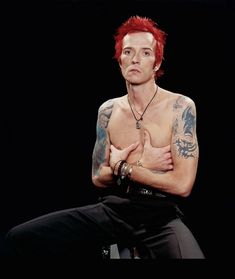 a man with red hair and tattoos on his chest sitting in front of a black background