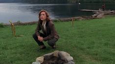 a woman kneeling down next to a fire pit on top of a lush green field