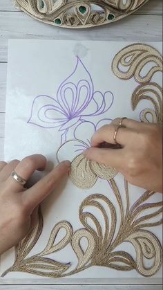 someone is working on some art work with gold and purple thread, while another person holds their hand over the paper