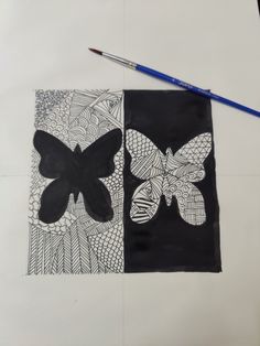 a drawing of two butterflies on black and white paper next to a blue pencil, with another butterfly in the background