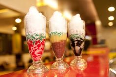 three glasses filled with ice cream and toppings sitting on a red counter top in a restaurant