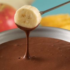 a banana being dipped into chocolate in a bowl with an apple and other fruit behind it