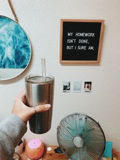 a person holding a cup in front of a desk with a fan and pictures on the wall