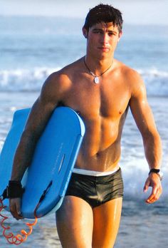 a shirtless man carrying a blue surfboard on the beach