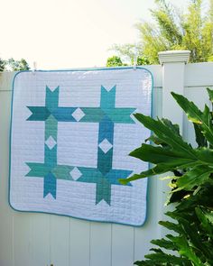 a quilted wall hanging on the side of a white fence next to a potted plant