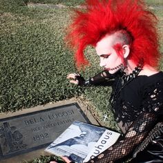 a woman with red hair sitting in the grass next to a plaque and holding a book