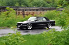 a black car driving down a wet road next to green bushes and trees in the background