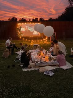 a group of people sitting around a table in the grass at night with balloons and lights