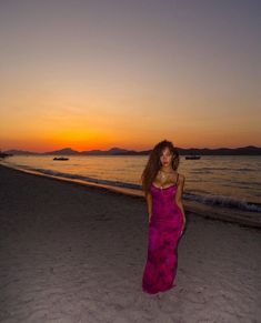 a woman in a pink dress standing on the beach at sunset with her hair blowing back