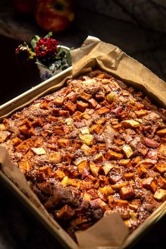 a baked dish in a cardboard box on a table next to an apple and flower arrangement