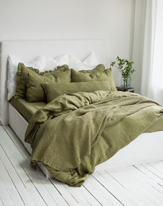 a bed with green sheets and pillows in a white room next to a plant on a table