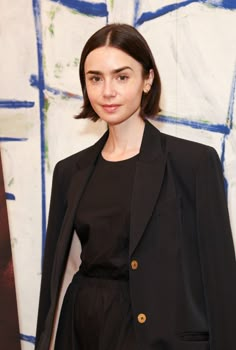 a woman standing in front of a painting wearing a blazer and black dress with gold buttons