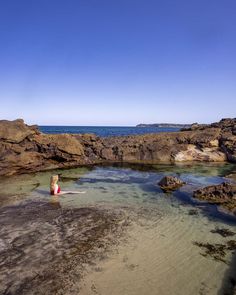 a woman sitting on top of a rock covered beach next to the ocean with her legs in the water