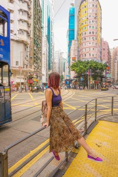 a woman standing on the side of a street next to a rail with buildings in the background