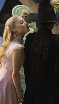 two women dressed in black and pink kissing each other with one woman's face obscured by her hat