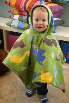 a little boy in a green dinosaur hooded blanket standing on the floor next to some pillows