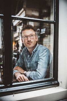 a man sitting at a window with his arms crossed and looking into the camera while wearing glasses