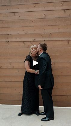 a man and woman standing next to each other in front of a wooden wall with their arms around each other