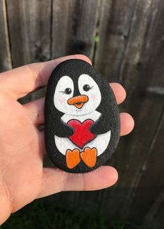 a hand holding a painted rock with a penguin on it's face and heart