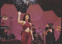 a woman in red outfit standing on stage with other people behind her and fireworks overhead