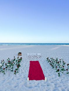 a red blanket on the beach with flowers in front of it that says happy valentine's day