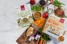 an assortment of dog food laid out on a marble counter top, including meats and vegetables