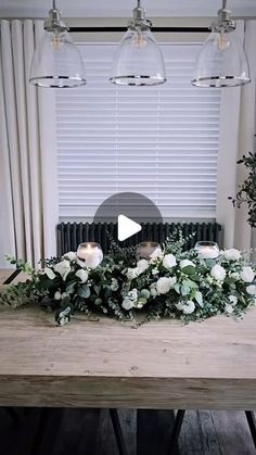 a wooden table topped with white flowers and greenery next to two hanging glass lights