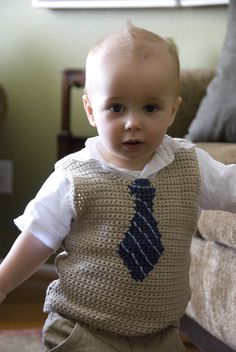 a little boy wearing a knitted vest and tie standing in front of a couch