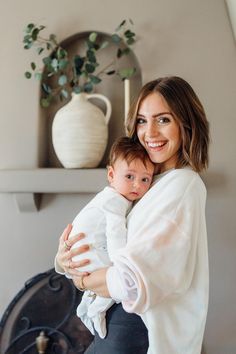 a woman holding a baby in her arms while standing next to a fireplace with a potted plant