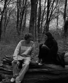 two people are sitting on a log in the woods, one is talking to the other