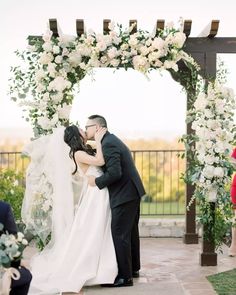 a bride and groom kissing under an arch decorated with white flowers at the end of their wedding ceremony