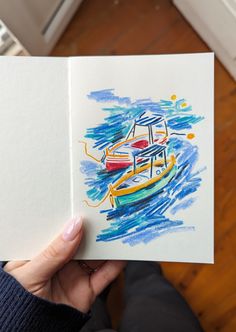 a person holding up an open book with watercolors on it and a boat in the background